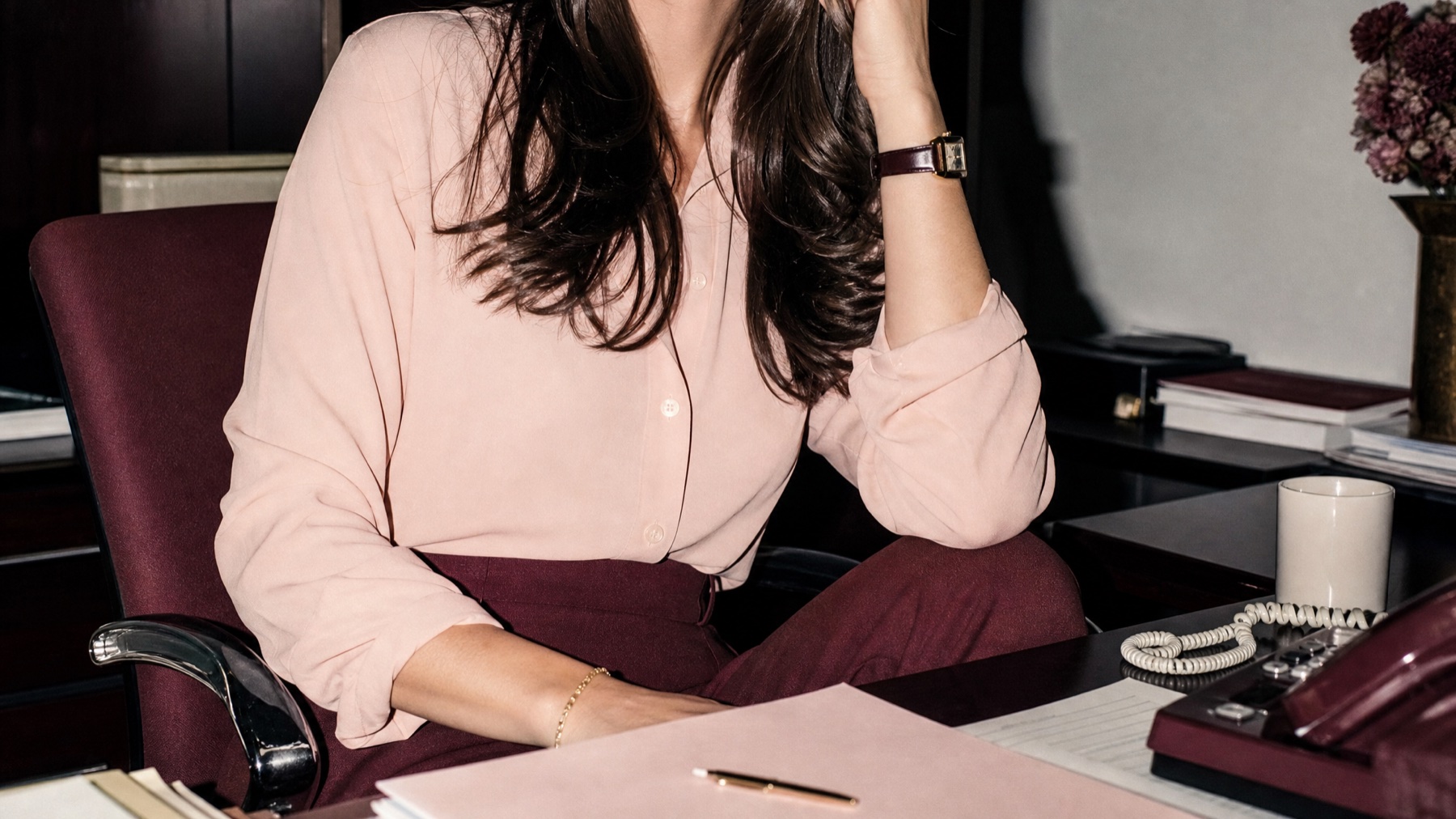 Candid photograph of a woman in a blush shirt and burgundy trousers at her desk